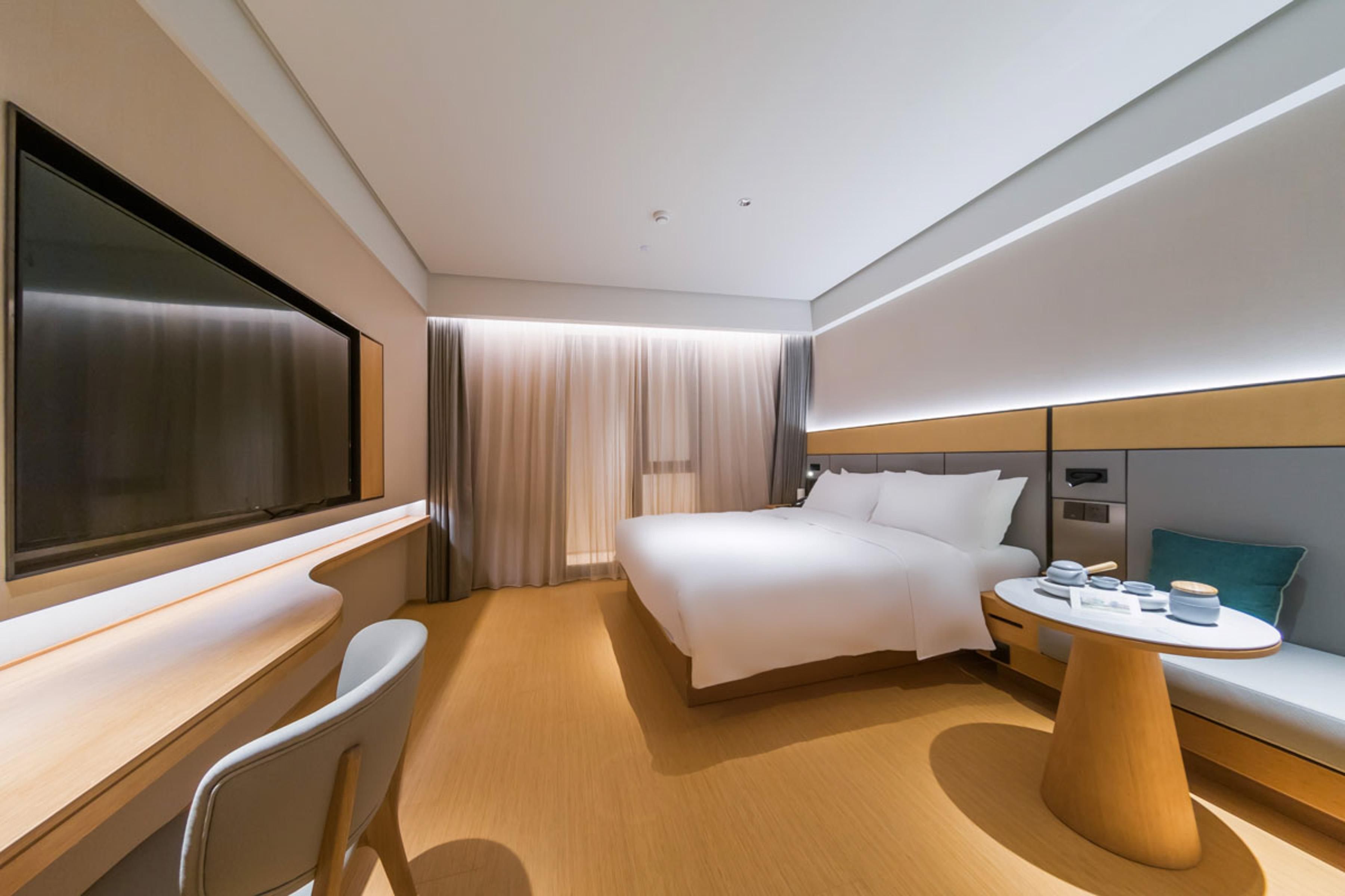 A modern hotel room with a large bed, a desk, a chair, a bedside table with a lamp, and a TV.
