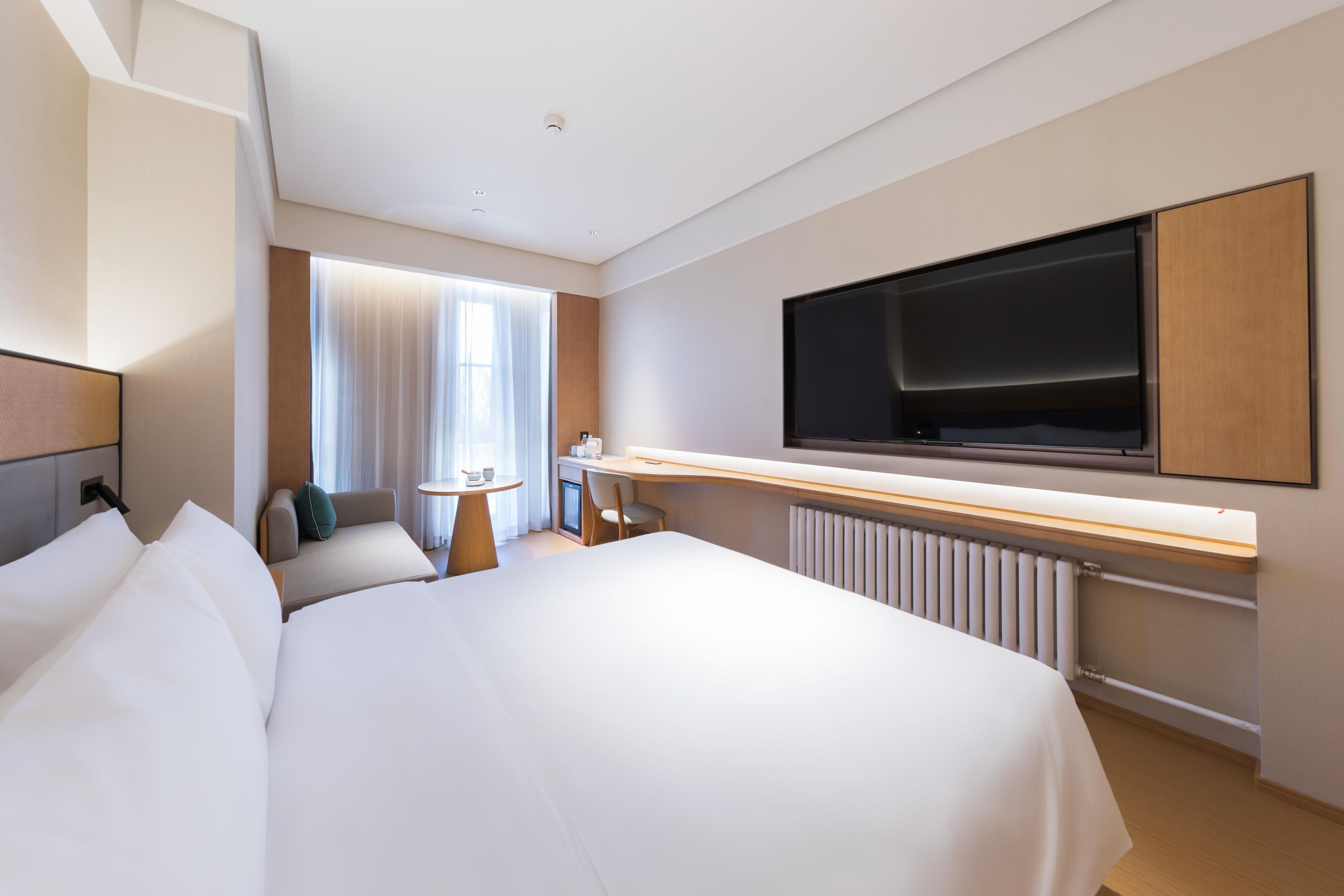 Een moderne hotelkamer met een groot bed, een aan de muur bevestigde televisie, een bureau met een stoel en een raam met gordijnen.