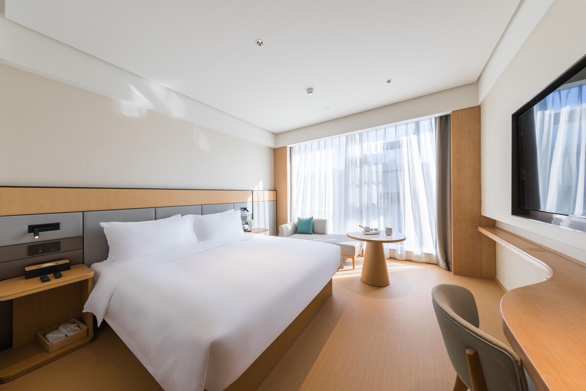 A hotel room with a large bed, bedside tables, a desk, a chair, a TV, and a window with curtains.