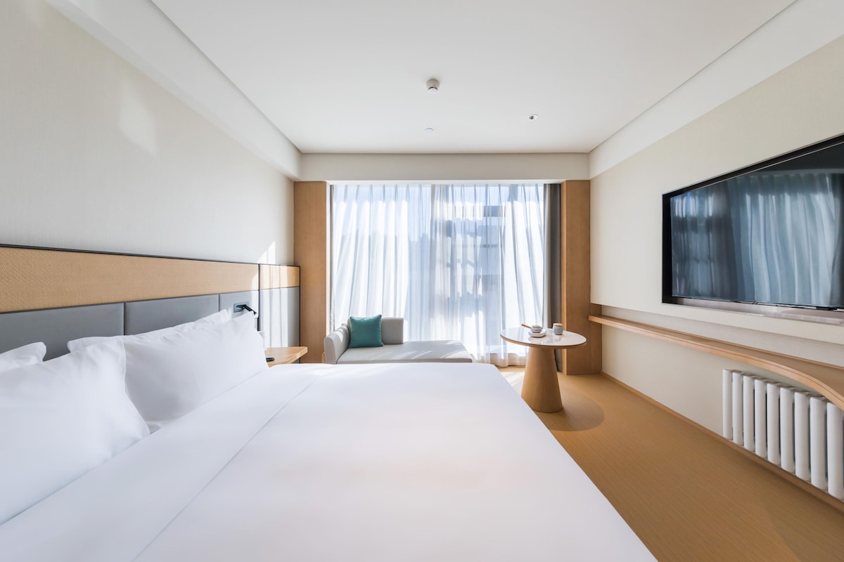 Ein Hotelzimmer mit einem großen Bett, einem Fernseher, einer Couch und einem Fenster mit Vorhängen.