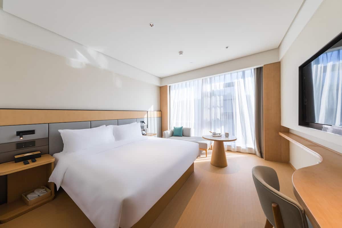 Ein Hotelzimmer mit einem großen Bett, Nachttischen, einem Schreibtisch, einem Sessel und einem Fernseher, sowie einem Fenster mit Vorhängen.