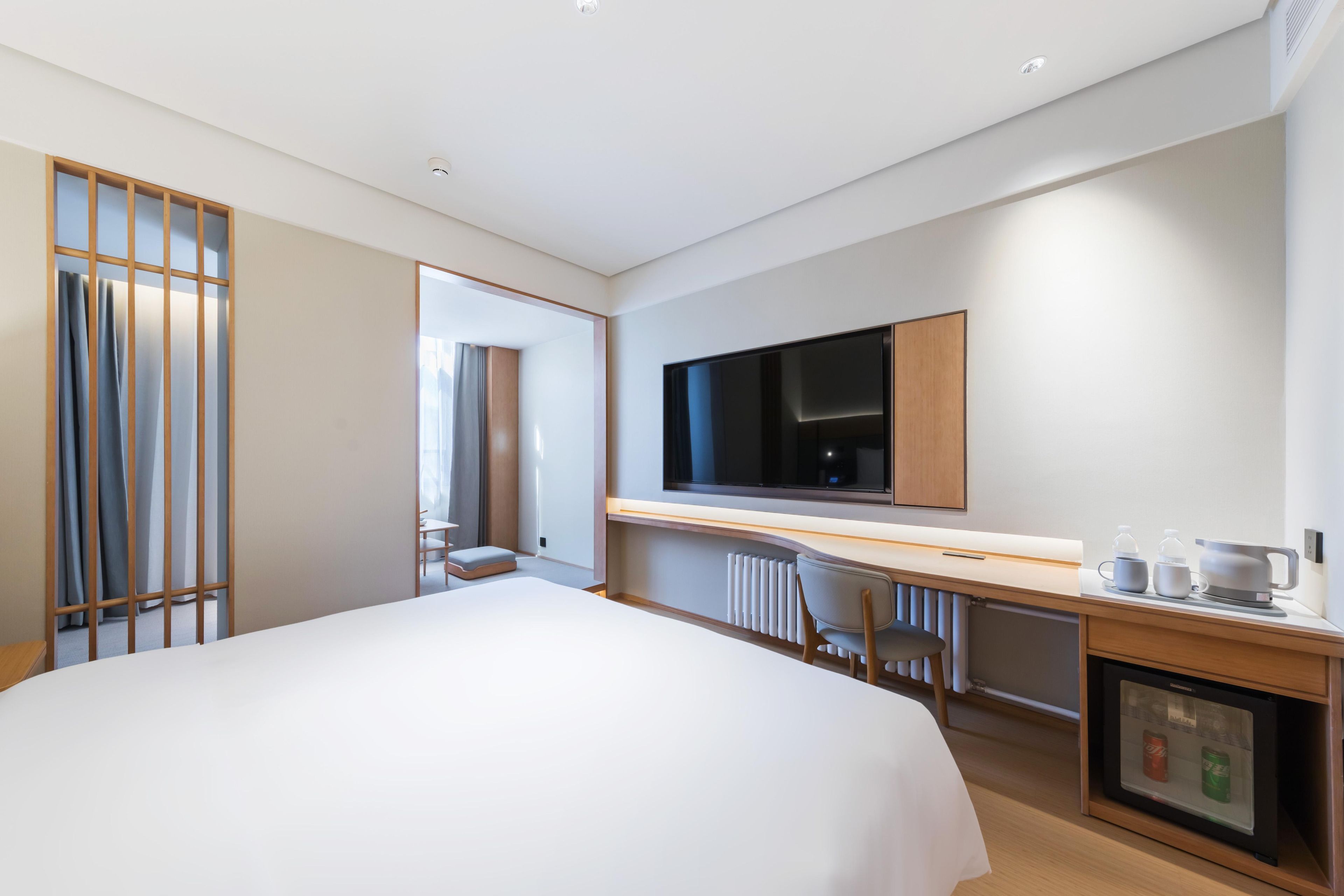 Ein modernes Hotelzimmer mit einem großen Bett, einem Fernseher, einem Schreibtisch mit Computer und einem kleinen Kühlschrank.