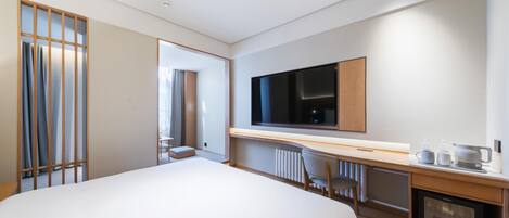 A modern hotel room with a large bed, a TV, a desk with a computer, and a mini-fridge.