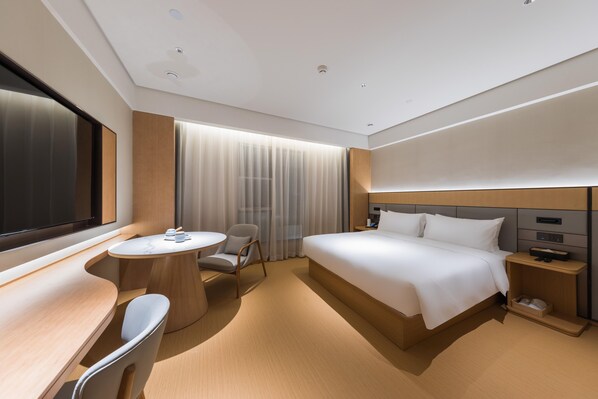 Quarto de hotel moderno com uma cama grande, uma escrivaninha com cadeira, uma pequena mesa redonda e uma janela ampla com cortinas.