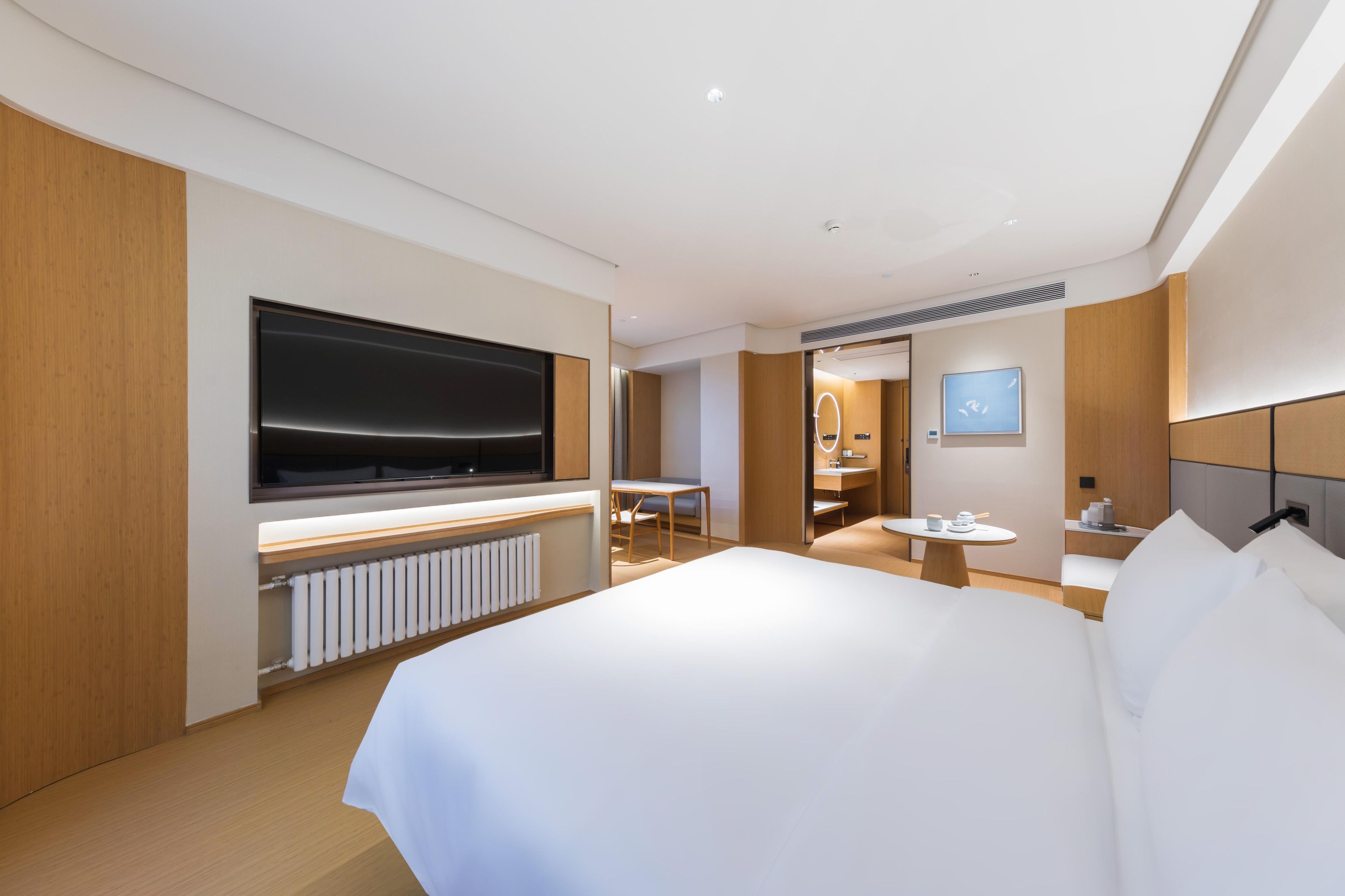 Una camera d'albergo moderna con un letto grande, una TV a schermo piatto fissata al muro, un piccolo tavolino con una sedia e un bagno visibile tramite una porta in vetro.