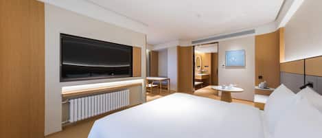 Una camera d'albergo moderna con un letto grande, una TV a schermo piatto fissata al muro, un piccolo tavolino con una sedia e un bagno visibile tramite una porta in vetro.
