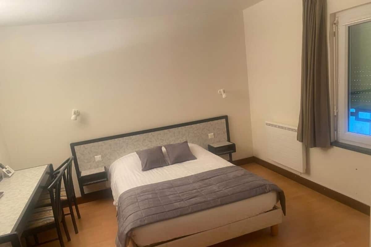 Un dormitorio con cama, mesita de noche, silla y ventana con cortinas.