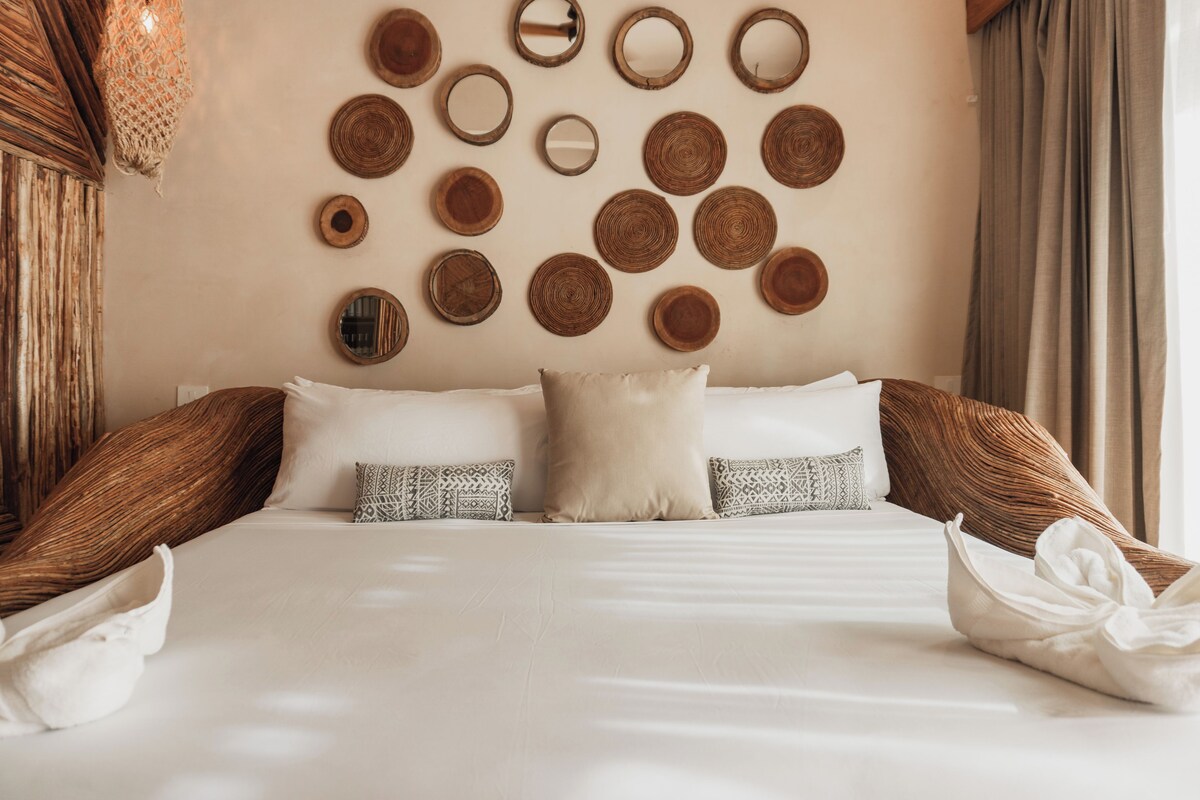 Quarto de hotel com uma cama bem arrumada, parede decorada com peças circulares trançadas e uma grande janela com cortinas.