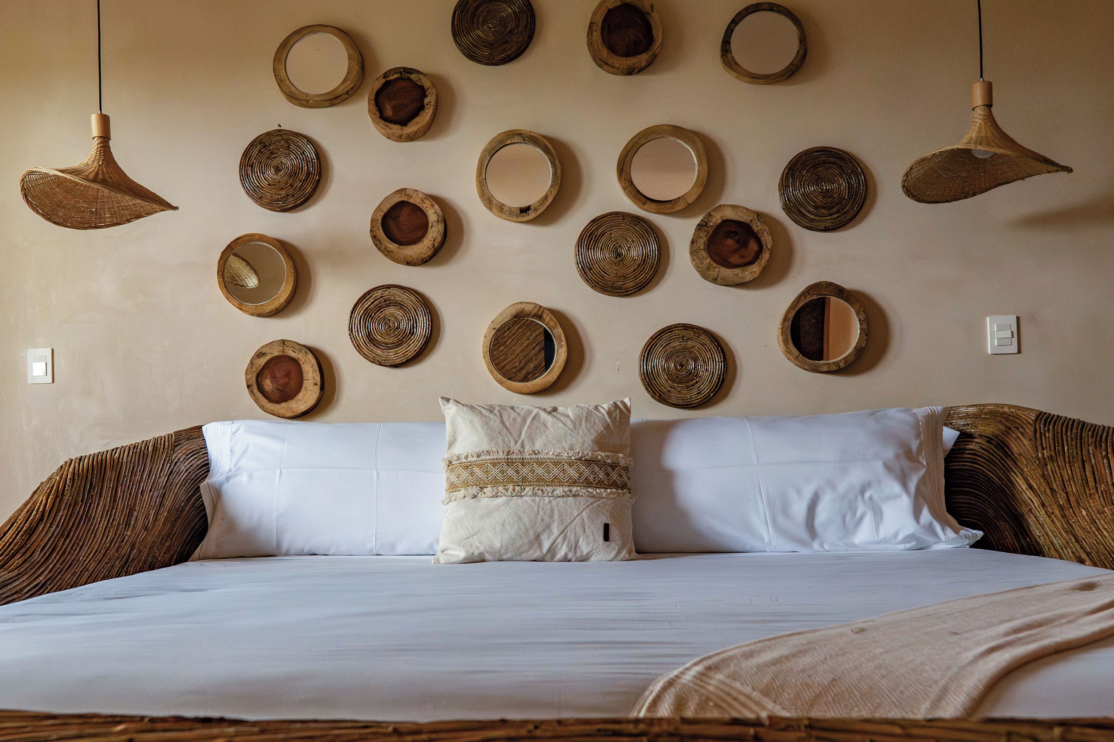 A bed with white linens and a patterned pillow, a wall with circular woven mirrors, and two hanging woven light fixtures.