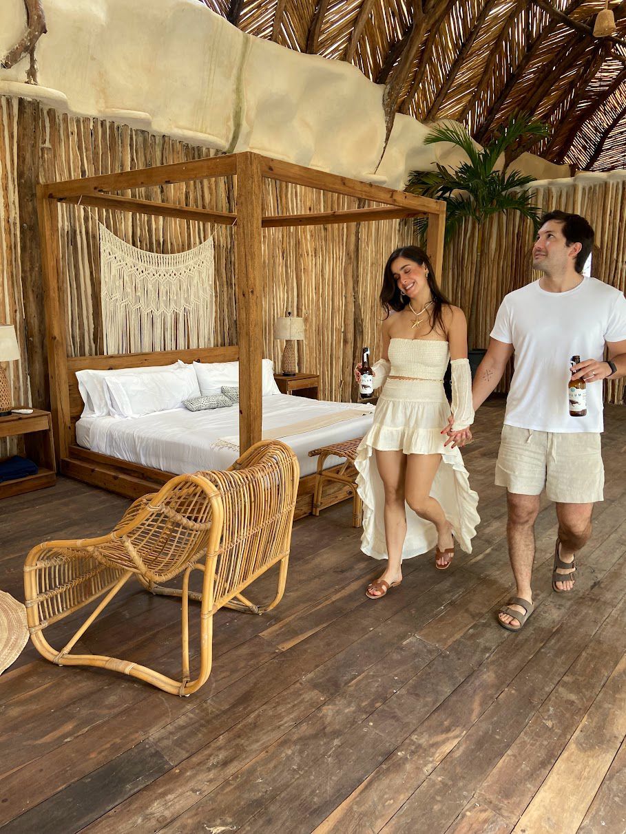 Una cama con dosel de madera con sábanas blancas, una silla de mimbre y un techo de paja tropical.
