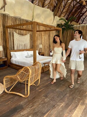 Uma cama de madeira com dossel e roupa de cama branca, uma cadeira de vime e um teto de palha tropical.