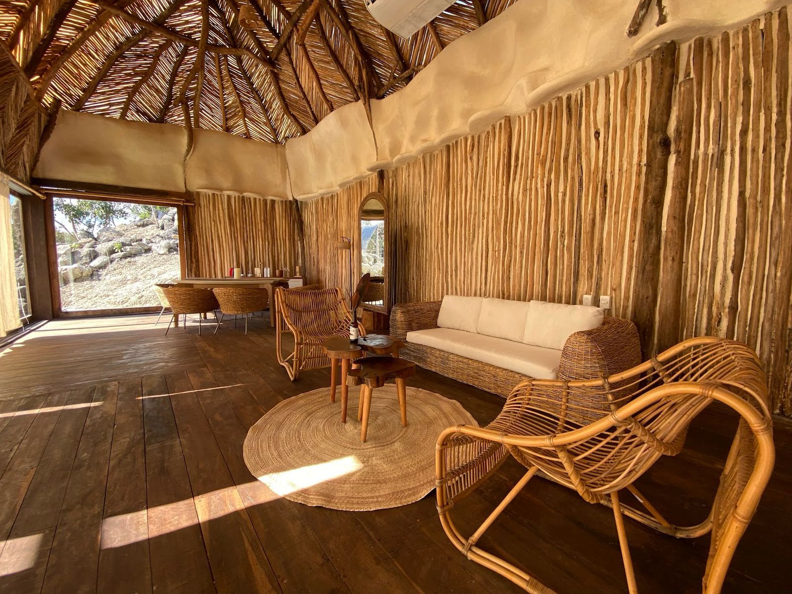 A room with a wooden floor, a sofa, wicker chairs, a round table, and a large window with a view of rocks.