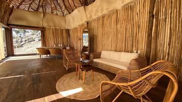 A room with a wooden floor, a sofa, wicker chairs, a round table, and a large window with a view of rocks.