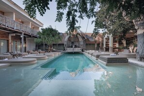 Uma piscina com espreguiçadeiras, uma estrutura com telhado de palha e um edifício moderno com varandas.