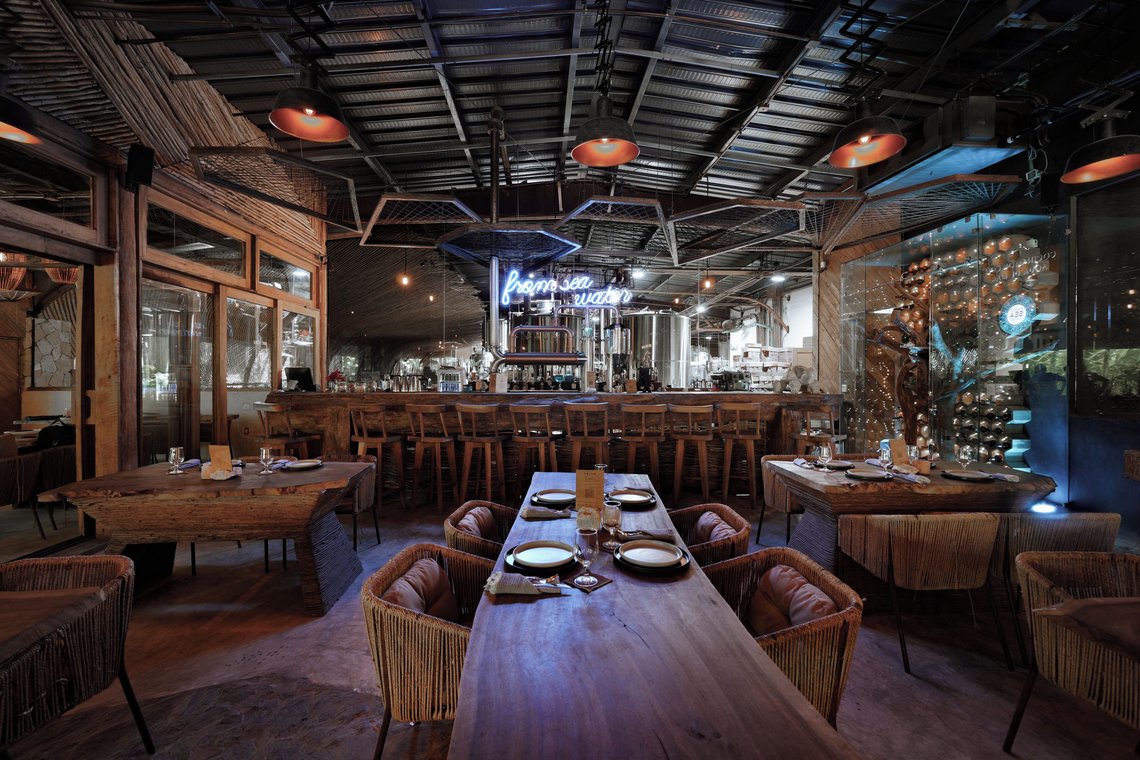 Un restaurante con una larga mesa de madera, sillas de mimbre y una zona de bar con taburetes.