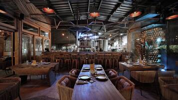 Un restaurante con una larga mesa de madera, sillas de mimbre y una zona de bar con taburetes.