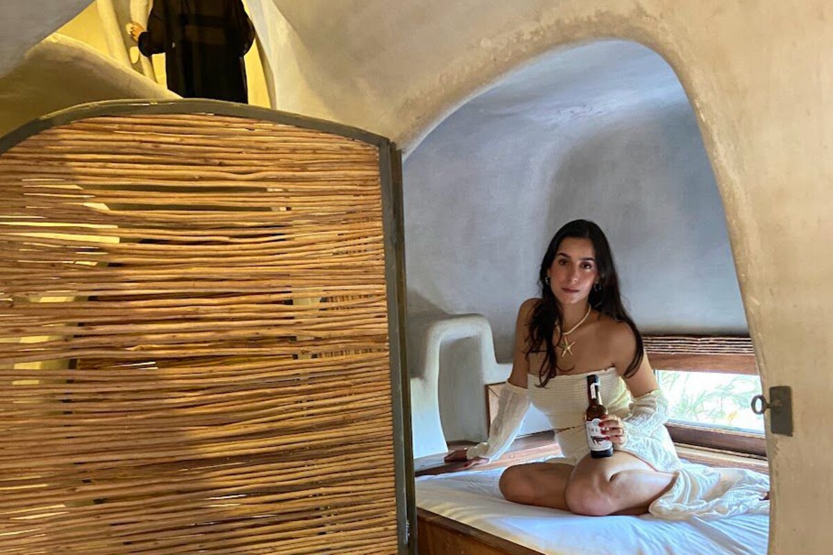 Una persona sentada en una cama dentro de una estructura en forma de cúpula geométrica con un muro de material similar al de la paja.