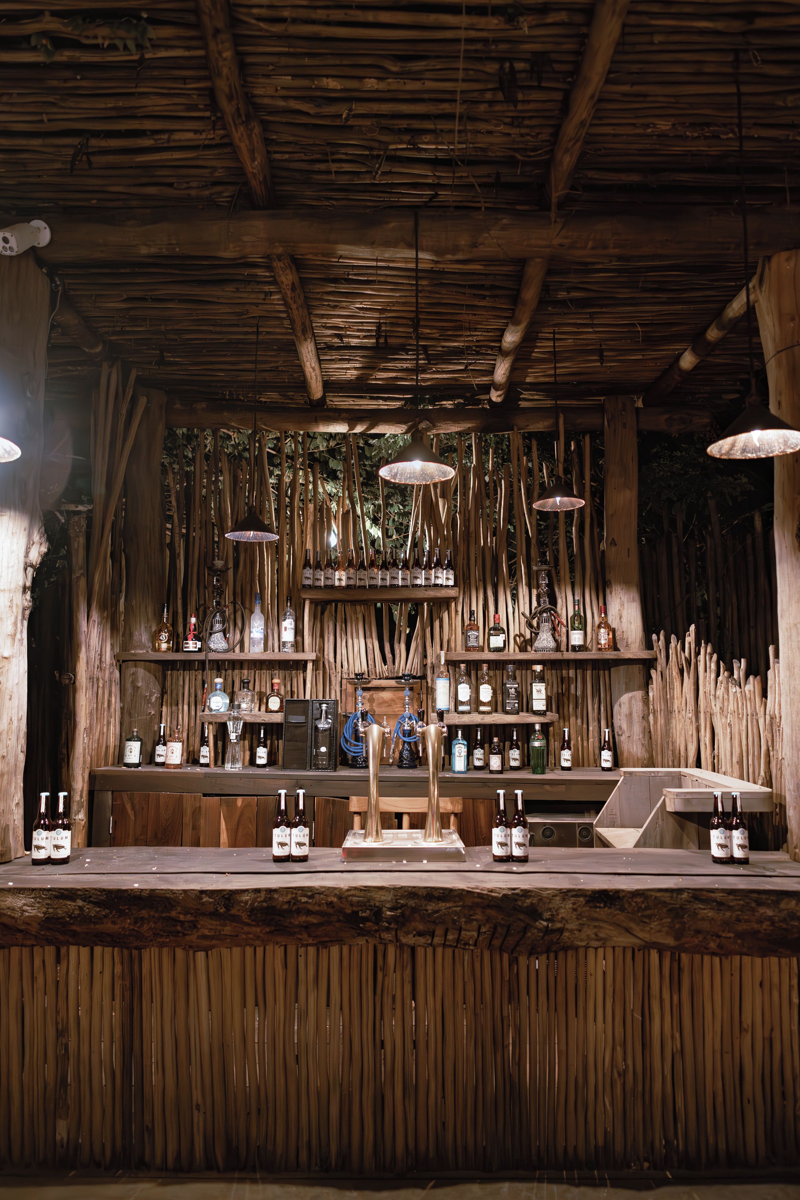 Un rústico área de bar con vigas de madera, luces colgantes y estantes llenos de botellas.