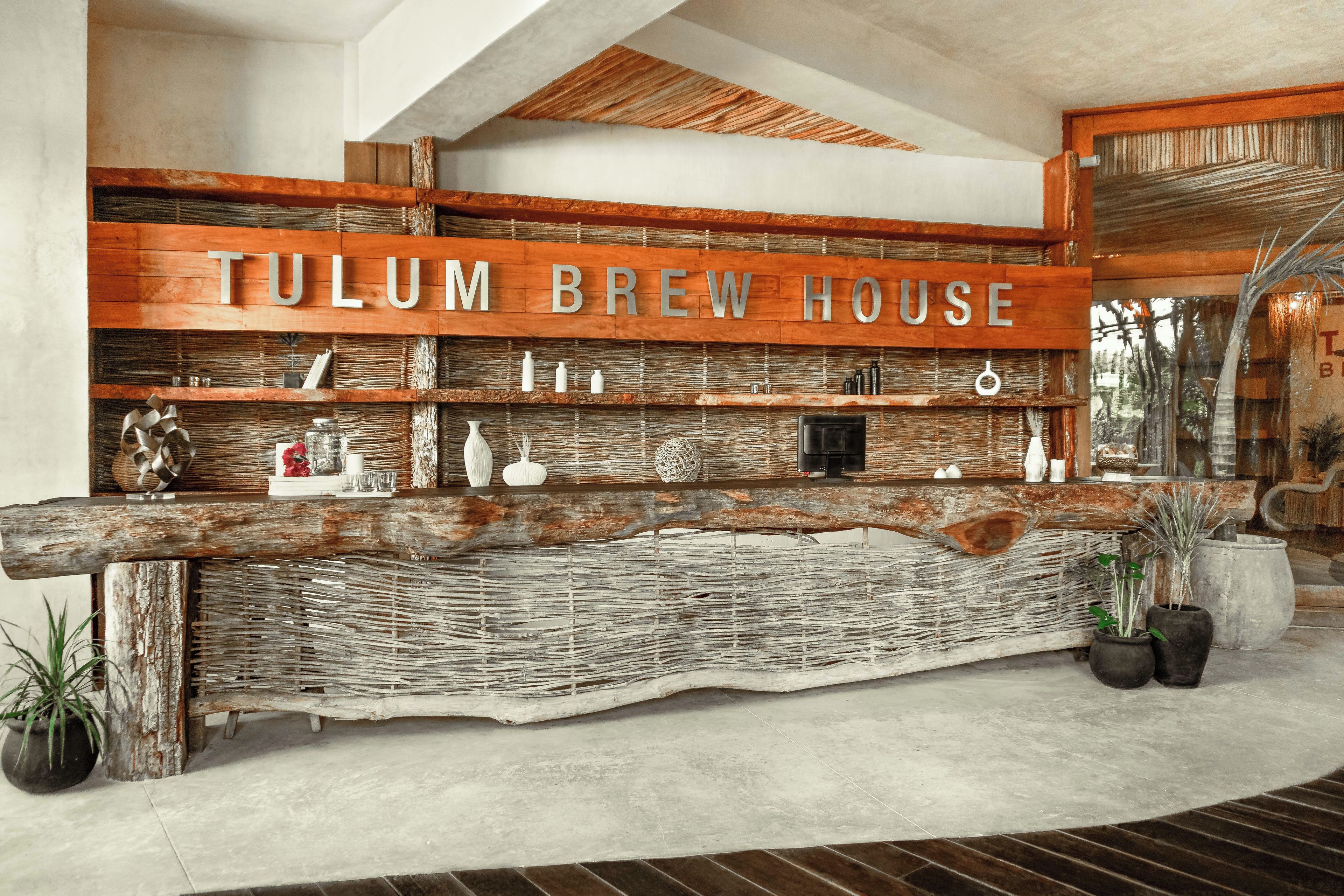Mostrador de recepción con letrero de 'TULUM BREW HOUSE', estantes tejidos y plantas en macetas.