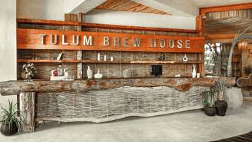Mostrador de recepción con letrero de 'TULUM BREW HOUSE', estantes tejidos y plantas en macetas.