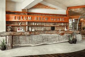 Mostrador de recepción con letrero de 'TULUM BREW HOUSE', estantes tejidos y plantas en macetas.