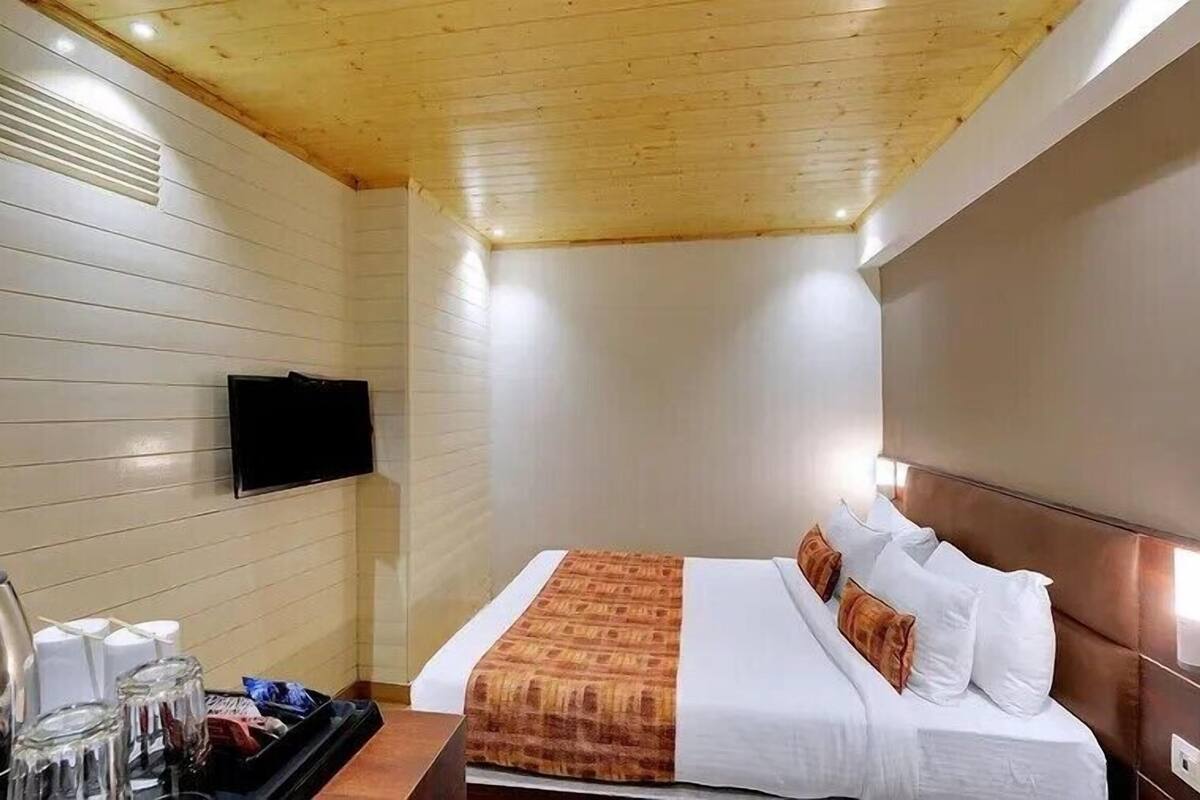 A hotel room with a bed, a TV mounted on the wall, and a small table with a kettle and glasses.