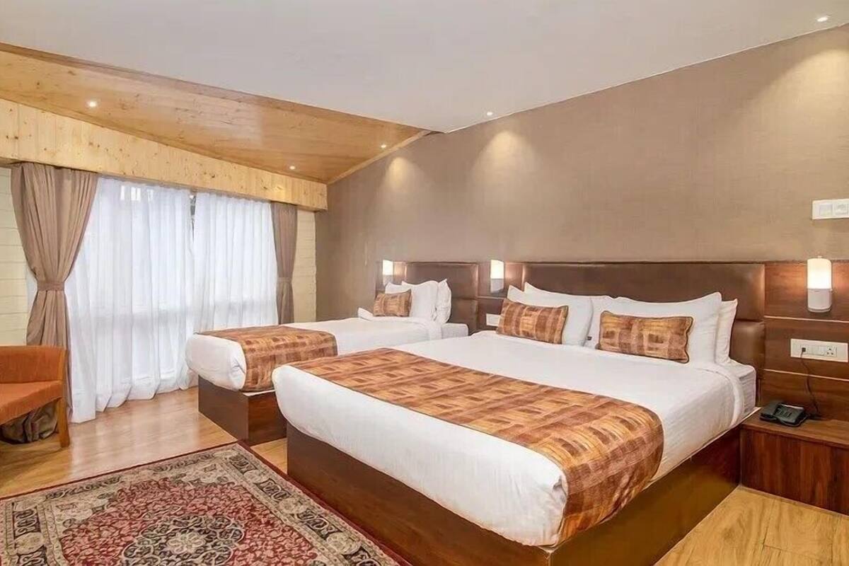 A hotel room with two beds, a wooden ceiling, a large window with curtains, a wooden chair, and a rug.