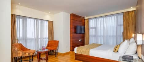 Una habitación de hotel con piso de madera, una cama con ropa blanca, un escritorio con silla y un televisor en la pared.