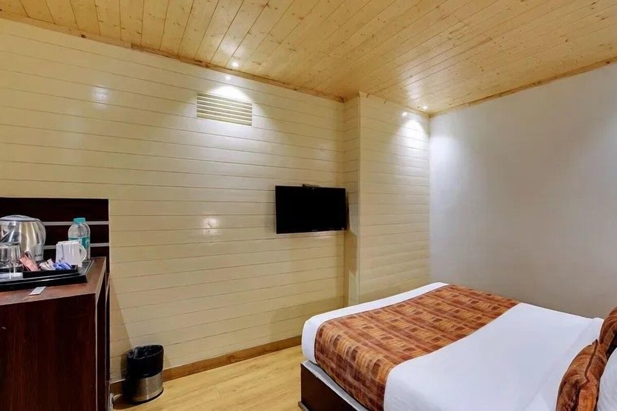 A hotel room with a wooden ceiling, a bed with a patterned blanket, a flat-screen TV mounted on the wall, and a small desk with a kettle and a water bottle.