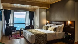 A hotel room with a wooden ceiling, a large bed with patterned bedding, a bedside table with a lamp, a chair, and a view of mountains through the window. - Sumitel Vista 800m From Mall Road (Darjeeling)