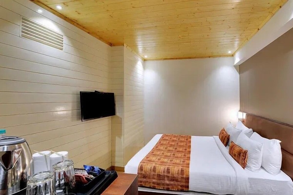 A hotel room with a bed, a TV mounted on the wall, and a small table with a kettle and glasses.