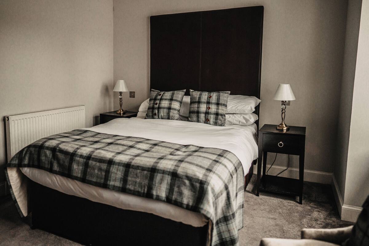 A neatly made bed with a plaid bedspread, two bedside tables with lamps, and a radiator on the wall.