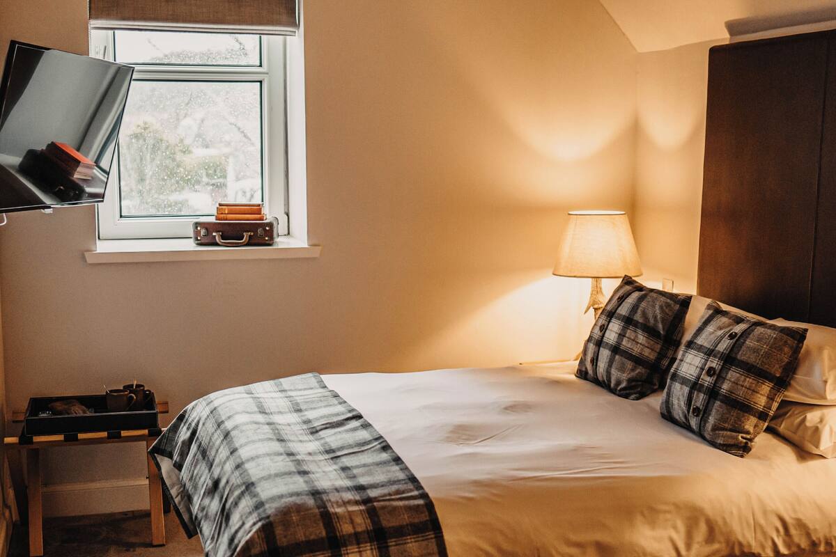 A neatly made bed with a plaid blanket and two pillows, a bedside table with a lamp, and a television on a stand.