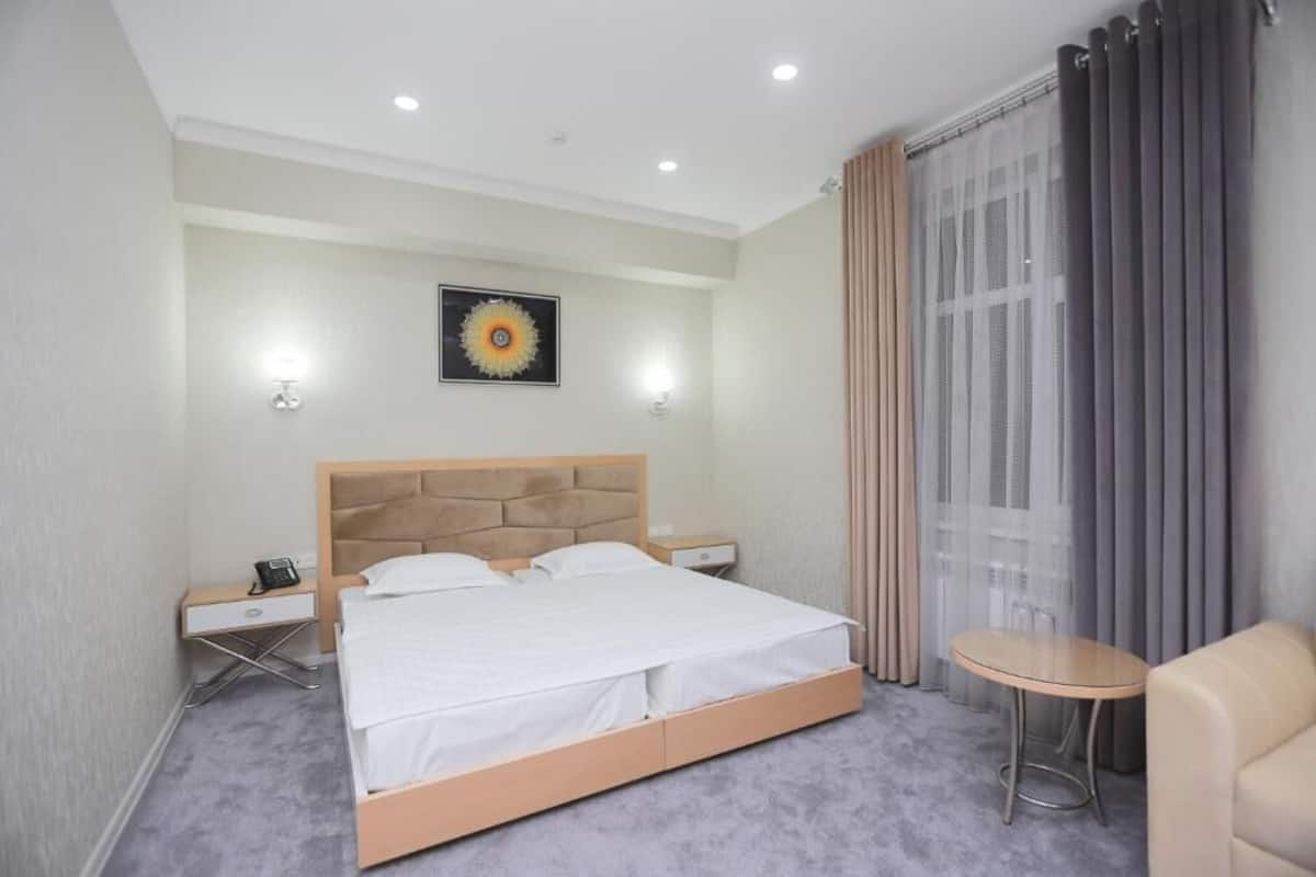 A hotel room with a bed, bedside tables, a small table, a chair, and a window with curtains.