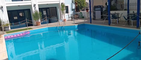 Una piscina con un flotador rosa, una zona de estar cubierta y un edificio con balcones.