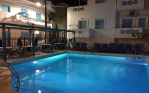 A swimming pool with lounge chairs and tables, surrounded by a building with balconies and windows.
