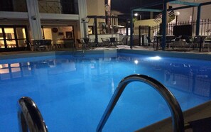 A swimming pool with a metal ladder, surrounded by chairs and tables, and a building with a balcony in the background.