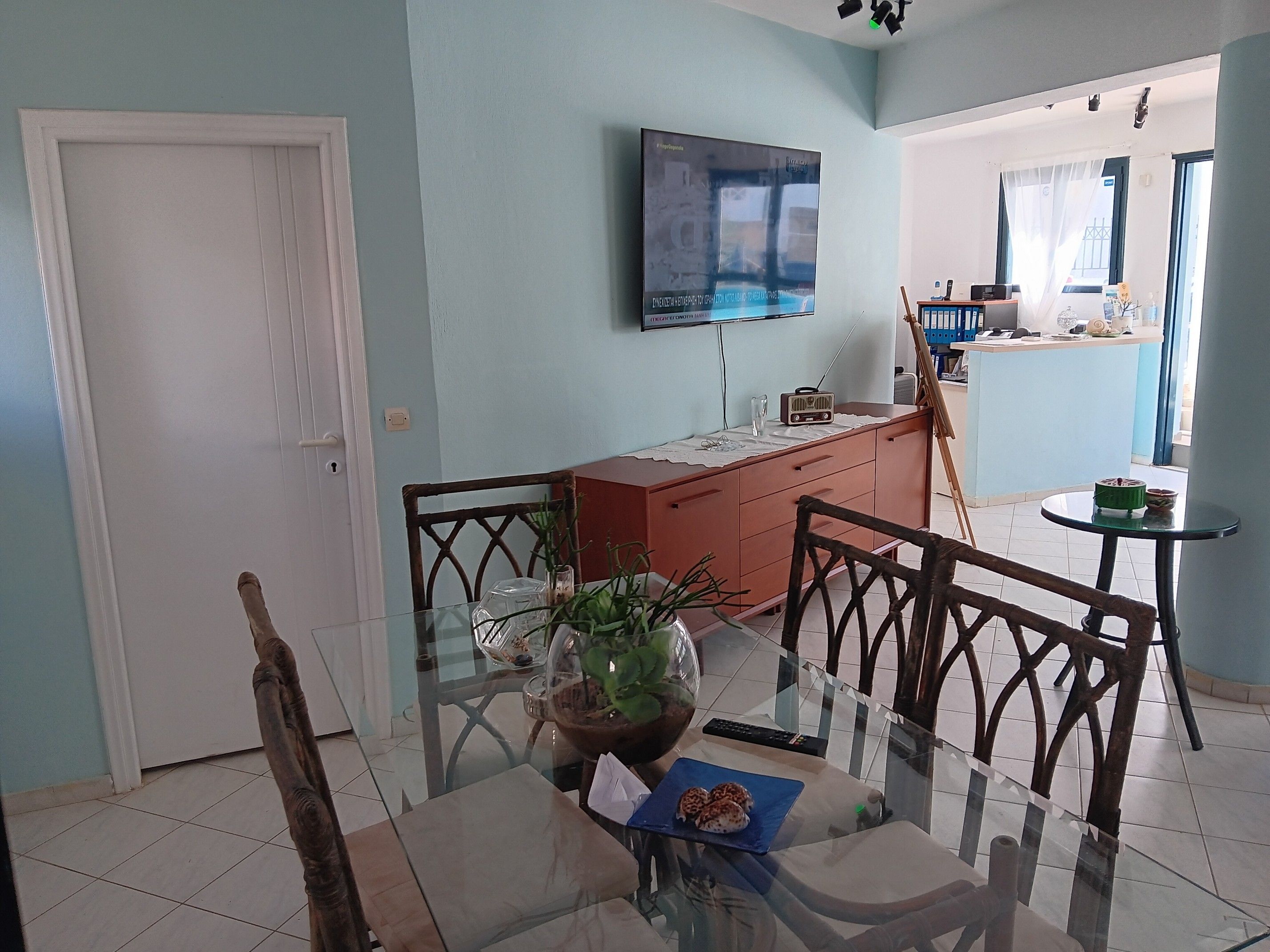 Uma sala de jantar com mesa de vidro, cadeiras, um vaso de planta e uma televisão na parede.