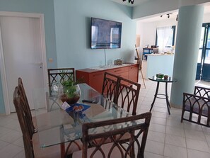 A dining area with a glass table, wooden chairs, a TV mounted on the wall, and a kitchen area in the background.