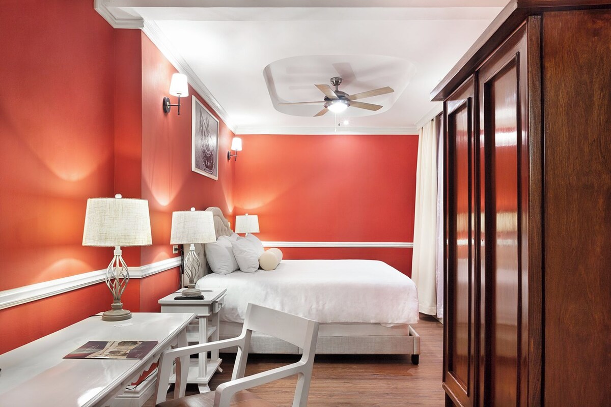 A hotel room with a bed, two bedside tables with lamps, a desk with a chair, a ceiling fan, and a large wooden wardrobe.