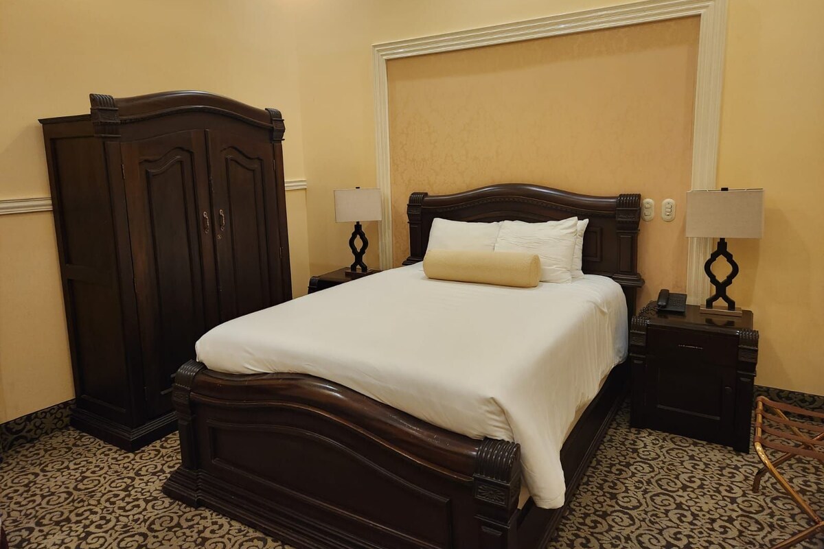 A neatly made bed with a wooden headboard, two bedside lamps, a dark wooden wardrobe, and patterned carpet.