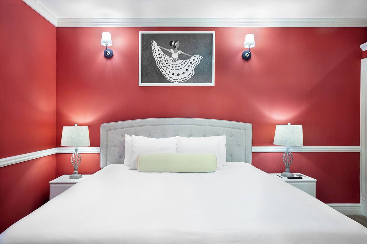 A hotel room with a large bed, two bedside lamps, a framed artwork above the bed, and red walls.