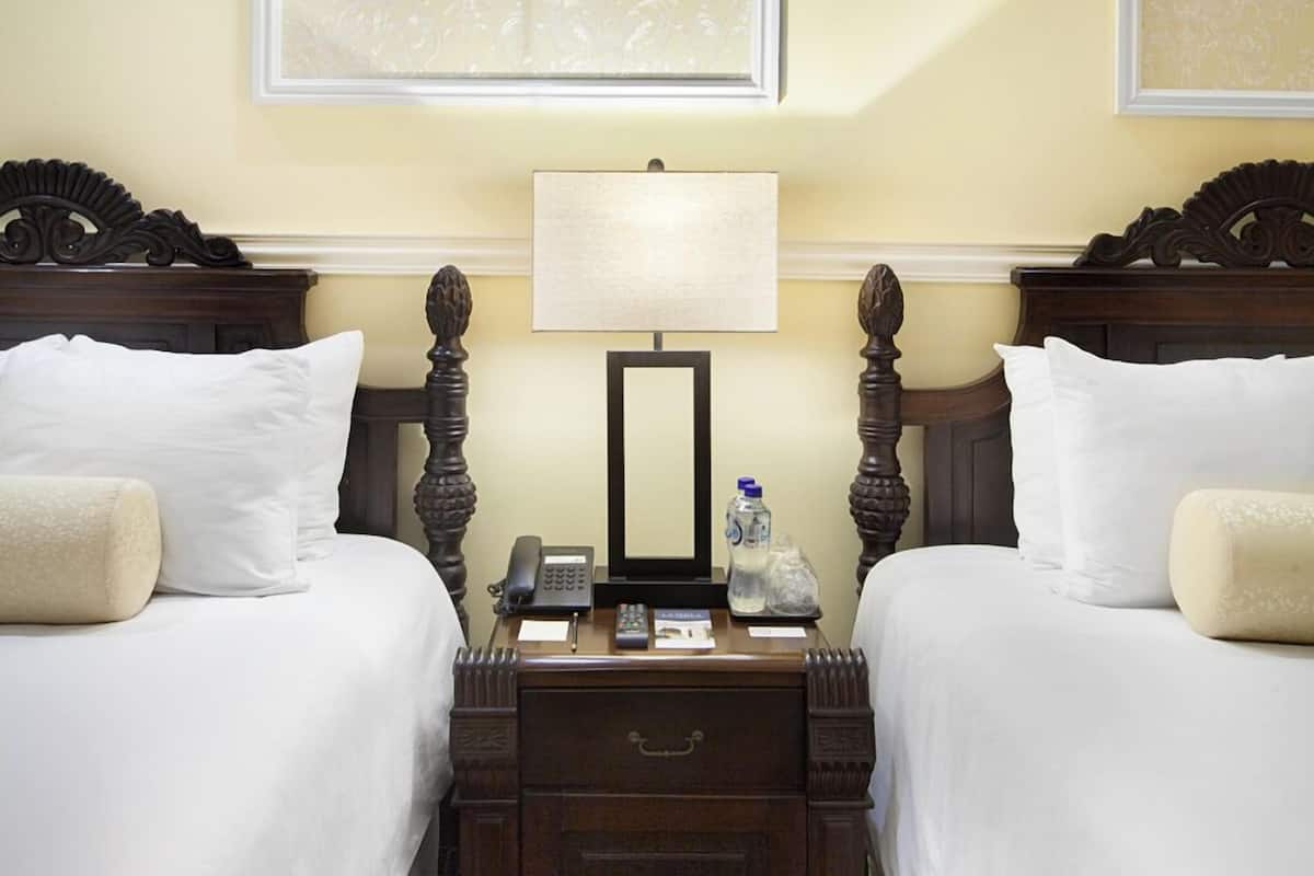 Habitación de hotel con dos camas, una mesita de noche con lámpara, teléfono y una botella de agua.