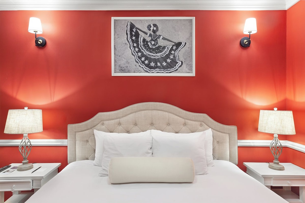 A hotel room with a red wall, a large bed with white linens, two bedside tables with lamps, and a framed artwork above the bed.