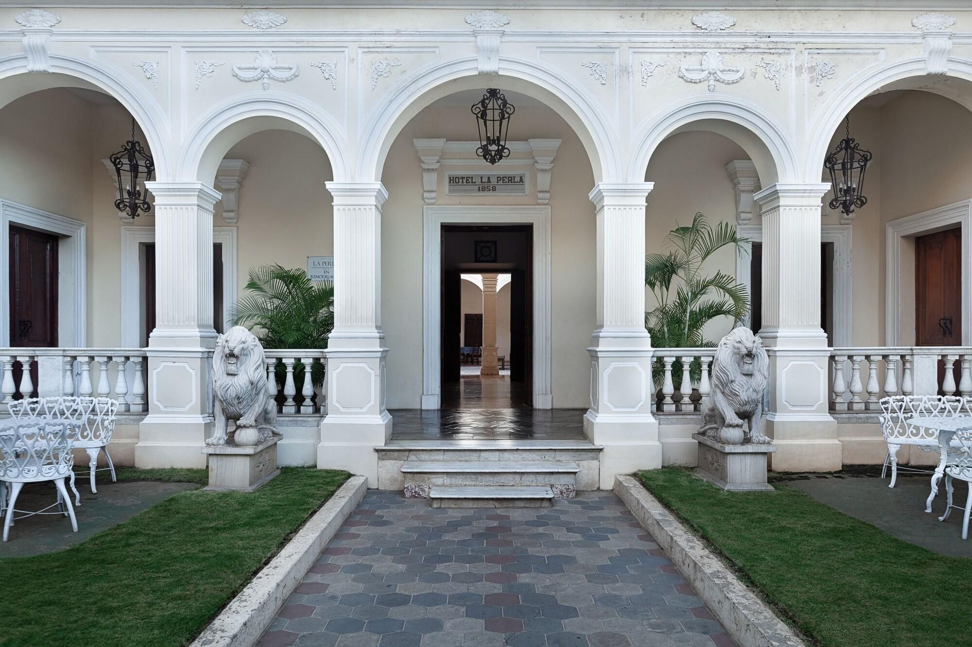 A historic building with a central entrance, flanked by white columns and ornate railings, leading to a covered walkway.