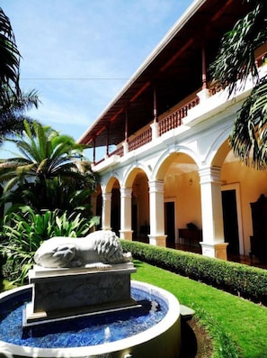 A building with a covered outdoor area, a fountain with a stone sculpture, and a well-maintained garden. - Hotel La Perla León (León)