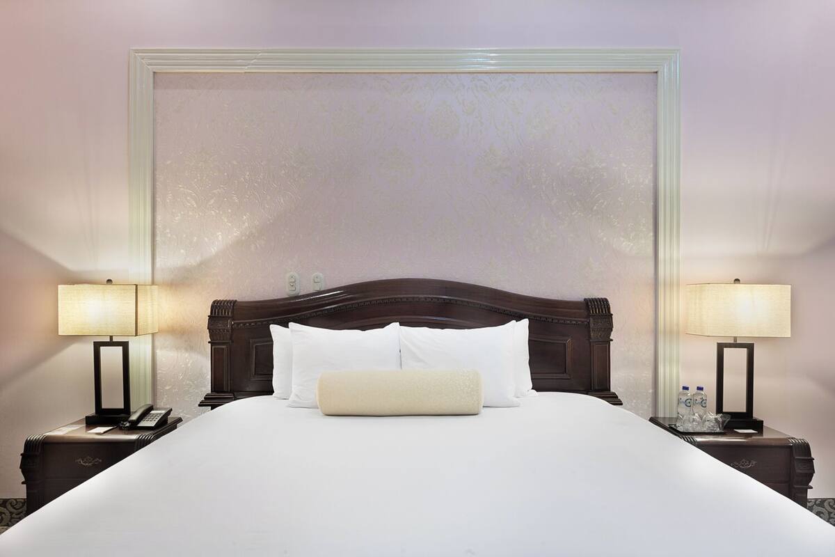A neatly made bed with white linens and a beige throw pillow, flanked by two bedside tables with lamps and a phone.