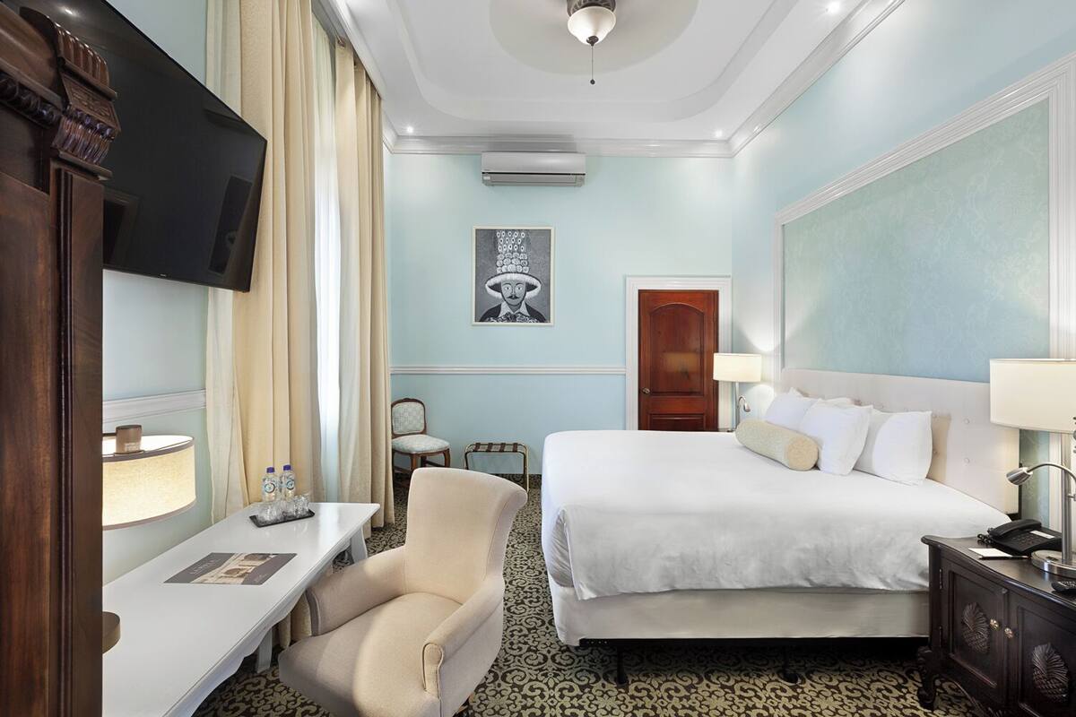 A hotel room with a bed, bedside table, chair, desk, TV, artwork, and an air conditioning unit.