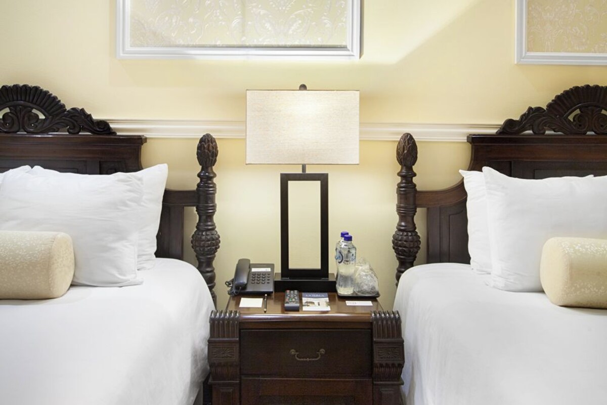 A hotel room with two beds, a nightstand, a lamp, and a mirror.