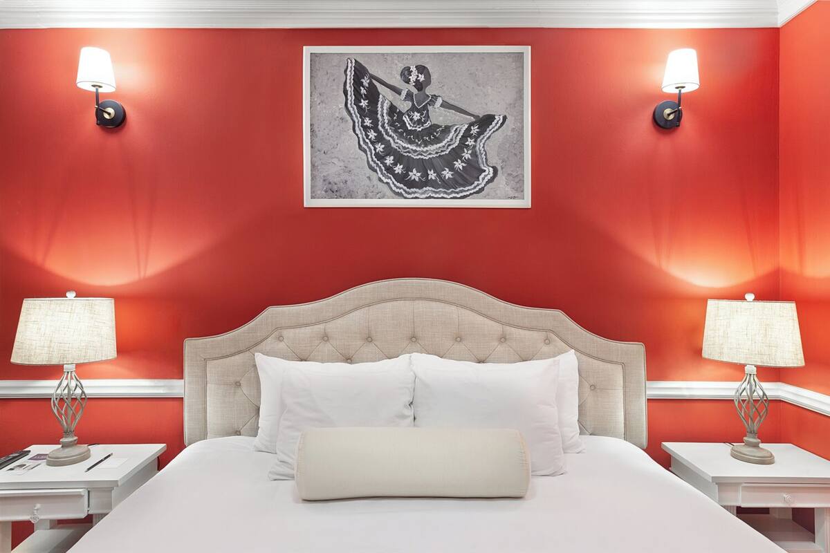 A hotel room with a red wall, a large bed with white linens, two bedside tables with lamps, and a framed artwork above the bed.